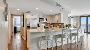 A modern kitchen with a central island, white bar stools, and a view of the ocean.