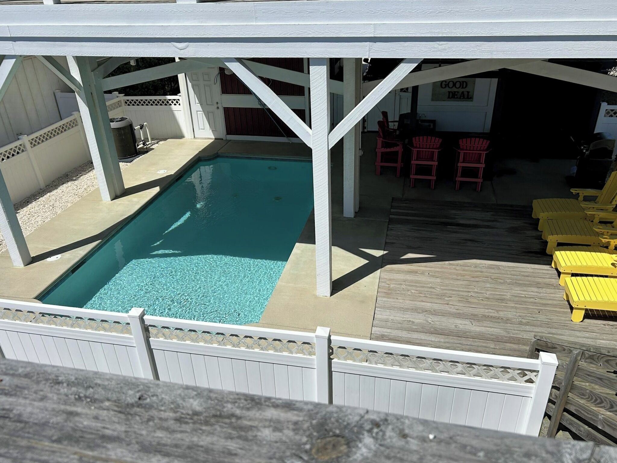 A rectangular pool with a white deck and a covered area with chairs.