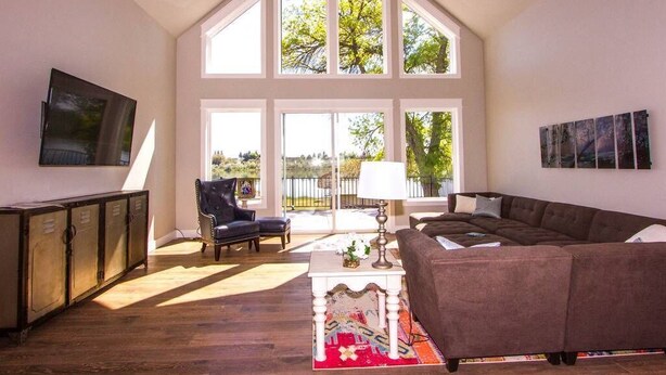 A spacious living room with a large window, a sofa, a chair, and a TV mounted on the wall. - Huge Lakefront House - sleeps 22! Private Dock! Boating, Water Skiing, Rafting! (Heyburn)