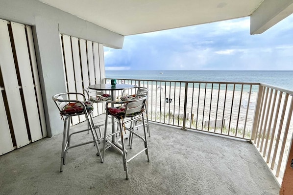 En balkon med metalstole og et bord, der giver udsigt over en strand med gelænder.
