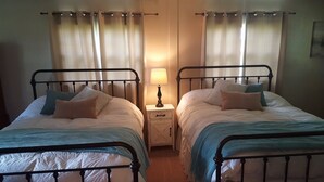 A room with two single beds, each with a nightstand, a lamp, and a window with curtains.
