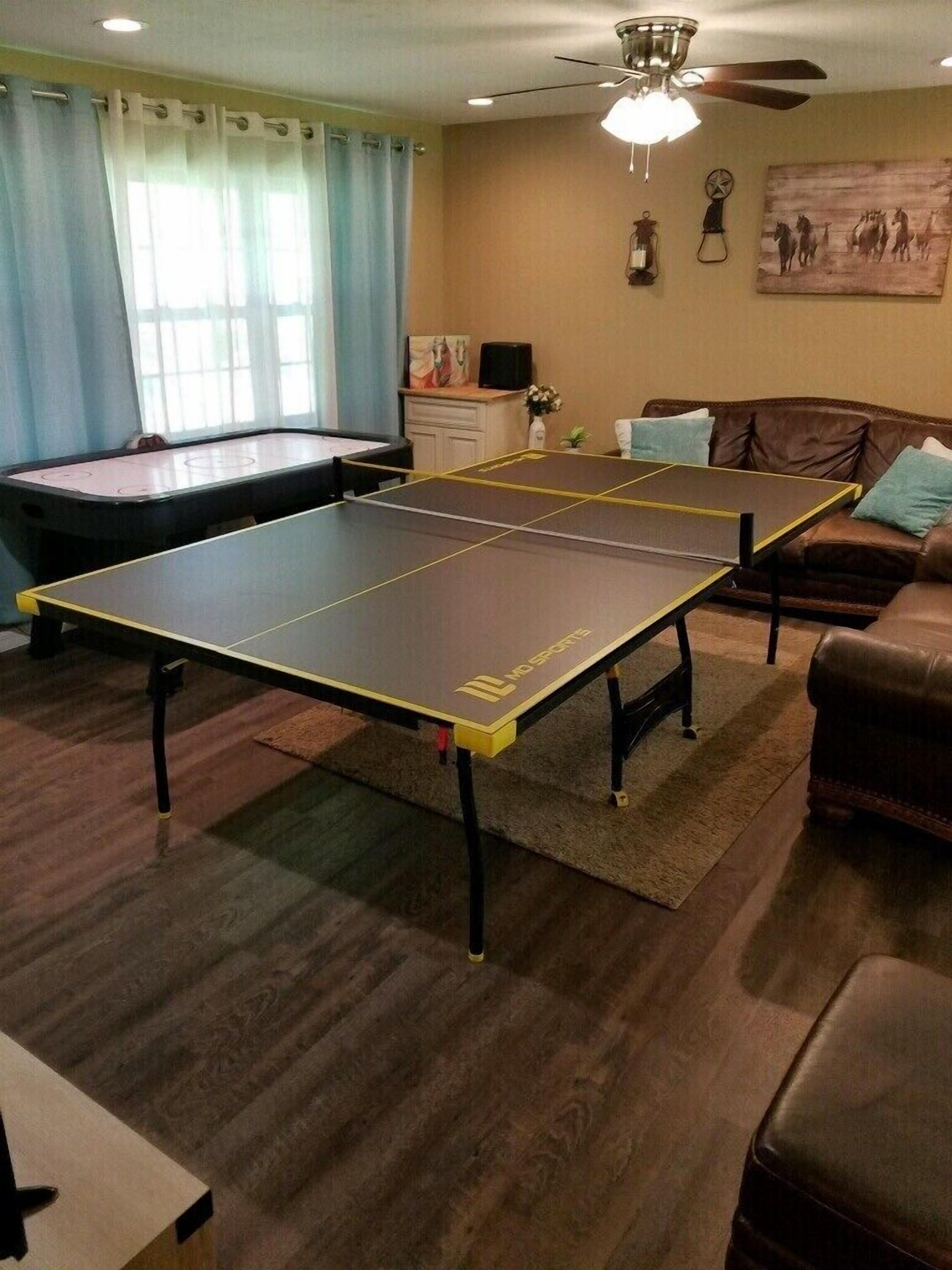A living room with a ping pong table, a sofa, a ceiling fan, and a painting on the wall.
