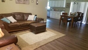 A living room with a brown leather sofa, a wooden coffee table, and a rug. There is a dining area with a wooden table and chairs. The kitchen is visible in the background with white cabinets.