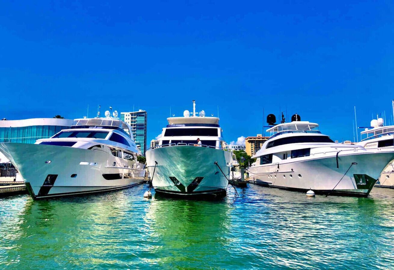 Three yachts docked at a marina with clear blue water and a city skyline in the background.