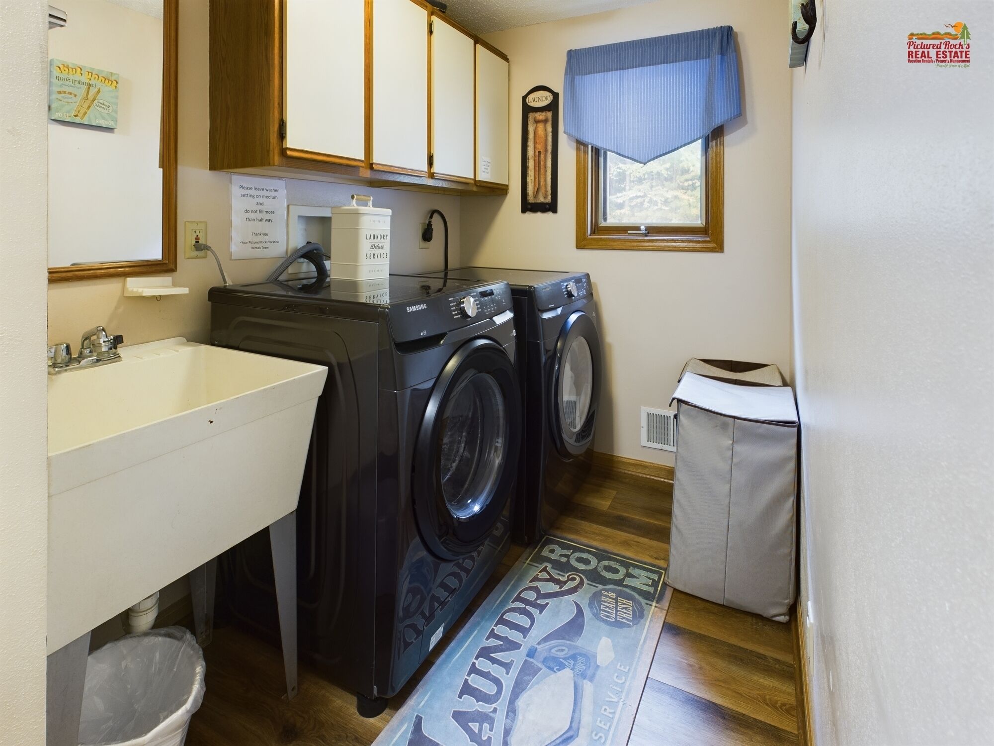 A laundry room with a sink, two washing machines, a trash can, and a laundry mat.