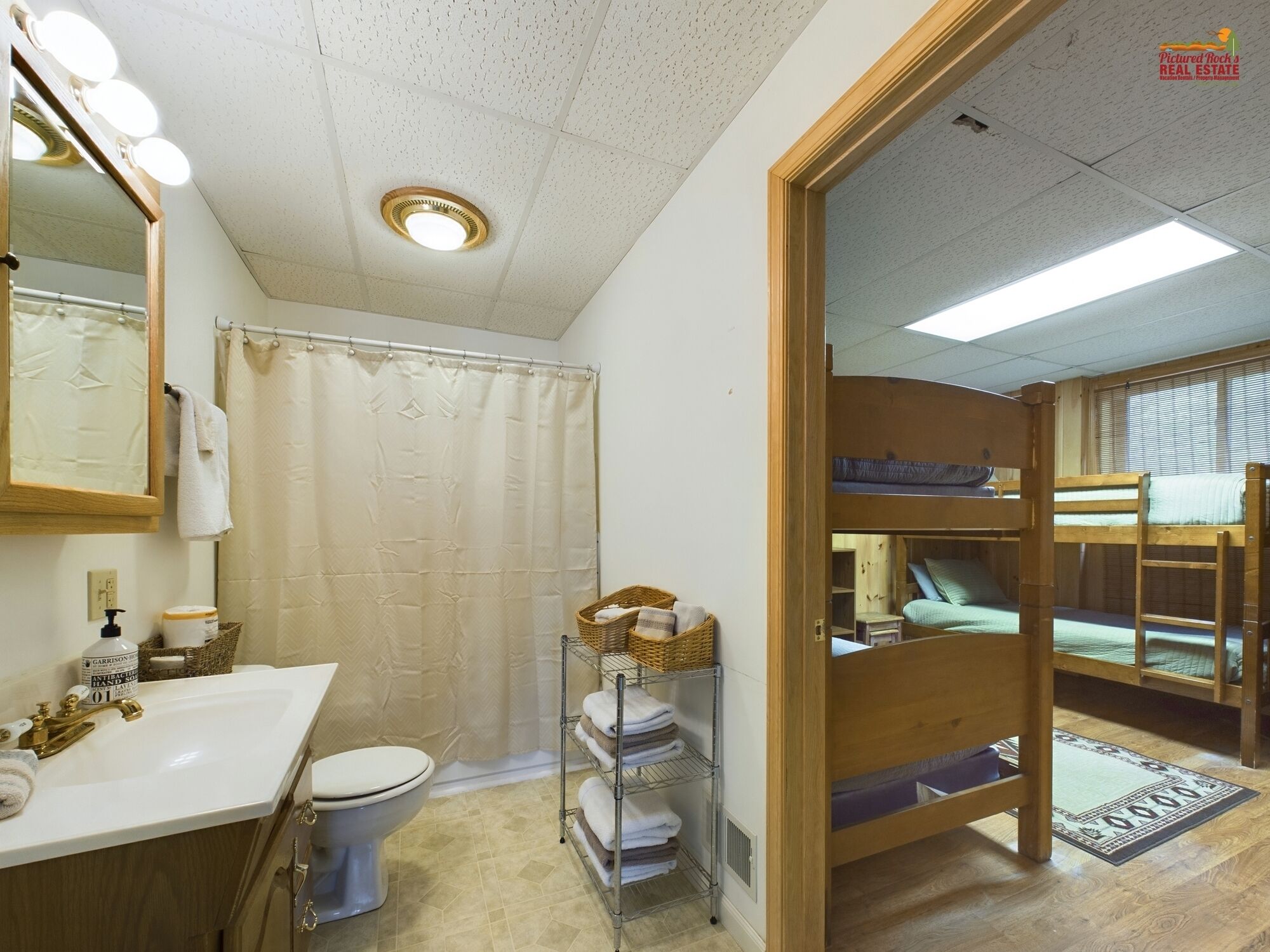 A bathroom with a sink, toilet, and shower curtain. There is a mirror with lights above it, and a wooden bunk bed in the adjacent room.