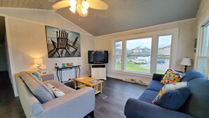 A living room with two sofas, a TV, a ceiling fan, and a window with a view of houses. - Barefootin' - steps from the beach, restaurants & shops. Very close to it all! (Kill Devil Hills)