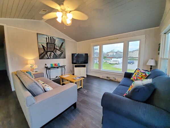 A living room with two sofas, a TV, a ceiling fan, and a window with a view of houses.