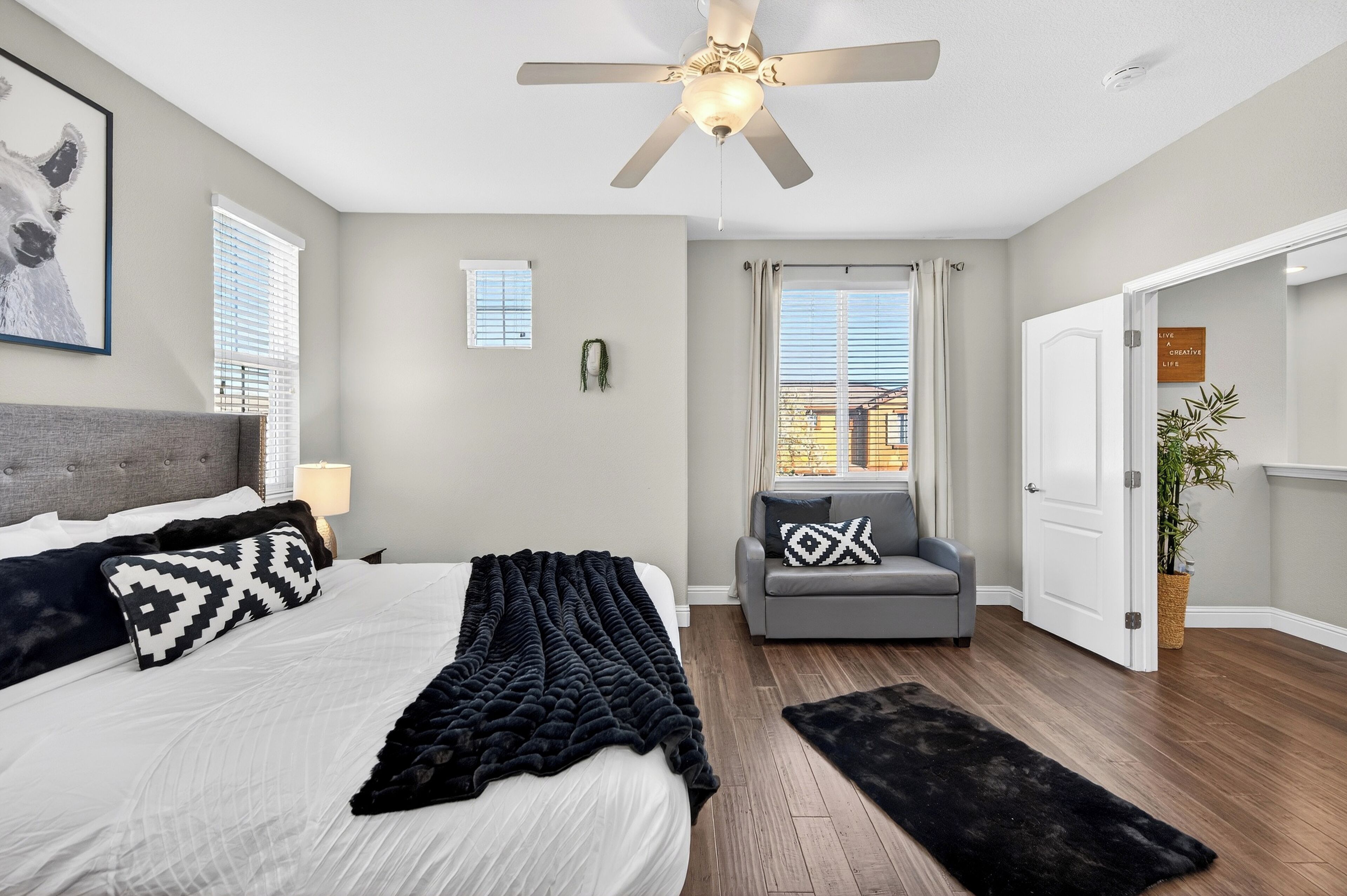 A bedroom with a bed, a gray sofa, a ceiling fan, a window with blinds, and a door leading to another room.