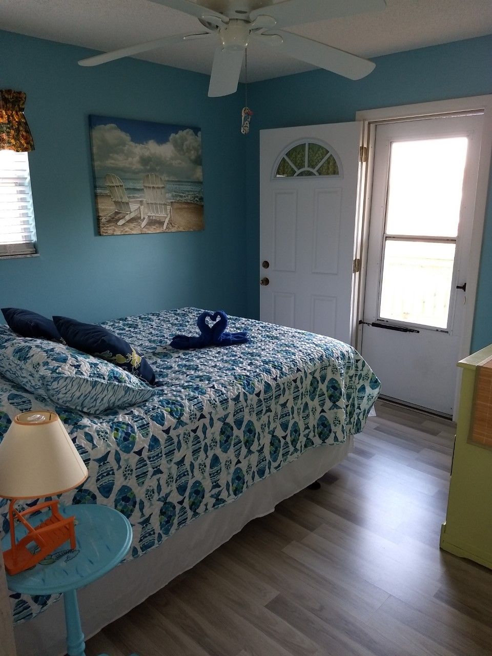A bedroom with a bed, a ceiling fan, a painting, a door, and a window.