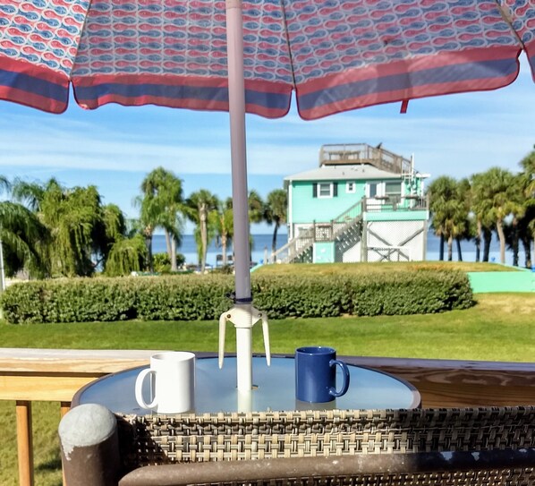 A table with a wicker chair, a white mug, and a blue mug under a patterned umbrella.
