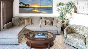 Ett vardagsrum med en beige soffa, ett soffbord i glas och en fåtölj med tropiskt mönster. En stor tavla som föreställer en solnedgång på stranden hänger på väggen.