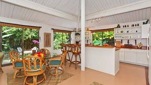 Una habitación amplia con comedor que incluye sillas de mimbre y una mesa de vidrio, una cocina con armarios blancos y una barra, y ventanas grandes con vistas a zonas verdes.