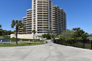 A tall, multi-story hotel with a modern design, featuring balconies on each floor, a paved walkway leading to the entrance, and a body of water in the background.