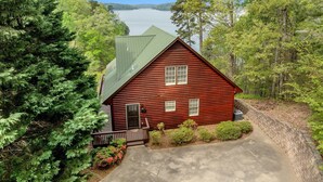 A red log cabin with a green roof, surrounded by dense forest and a lake in the background. - Log Cabin On Lake Keowee-Big Lake Mountain Views (Six Mile)
