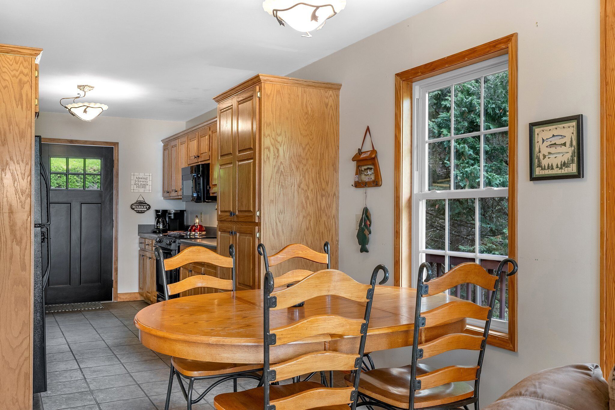A dining area with a wooden table and chairs, a kitchen in the background, and a window with a view of trees.