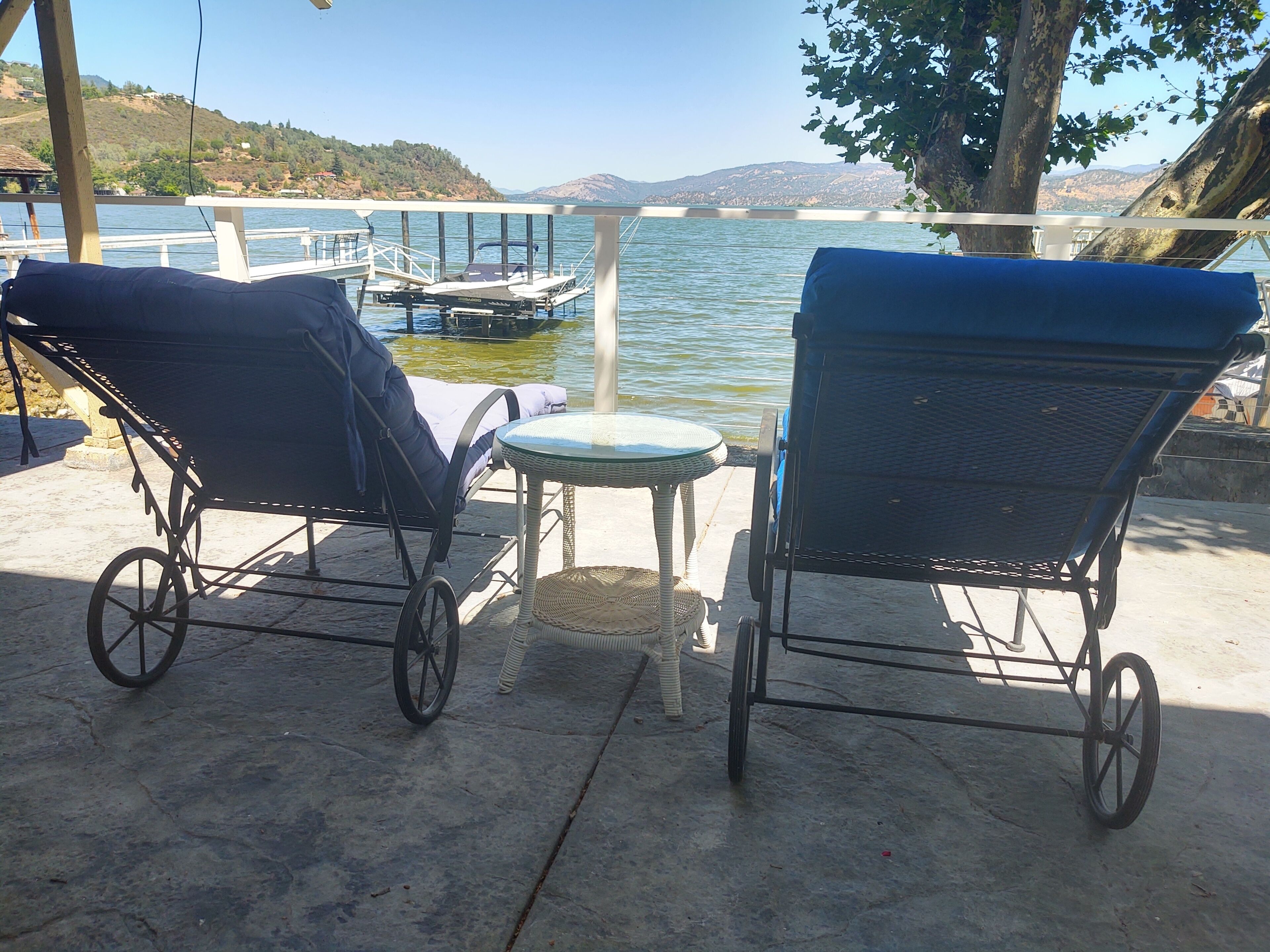 Deux fauteuils de jardin avec roulettes, une petite table ronde et une vue sur un lac, avec des bateaux et des collines en arrière-plan.