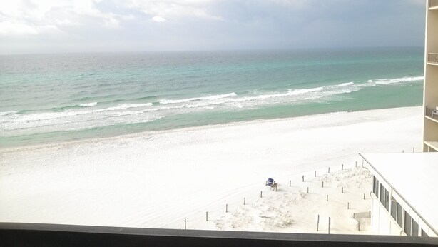A beach view from a balcony with a clear sky, a sandy beach, and a few chairs and umbrellas.
