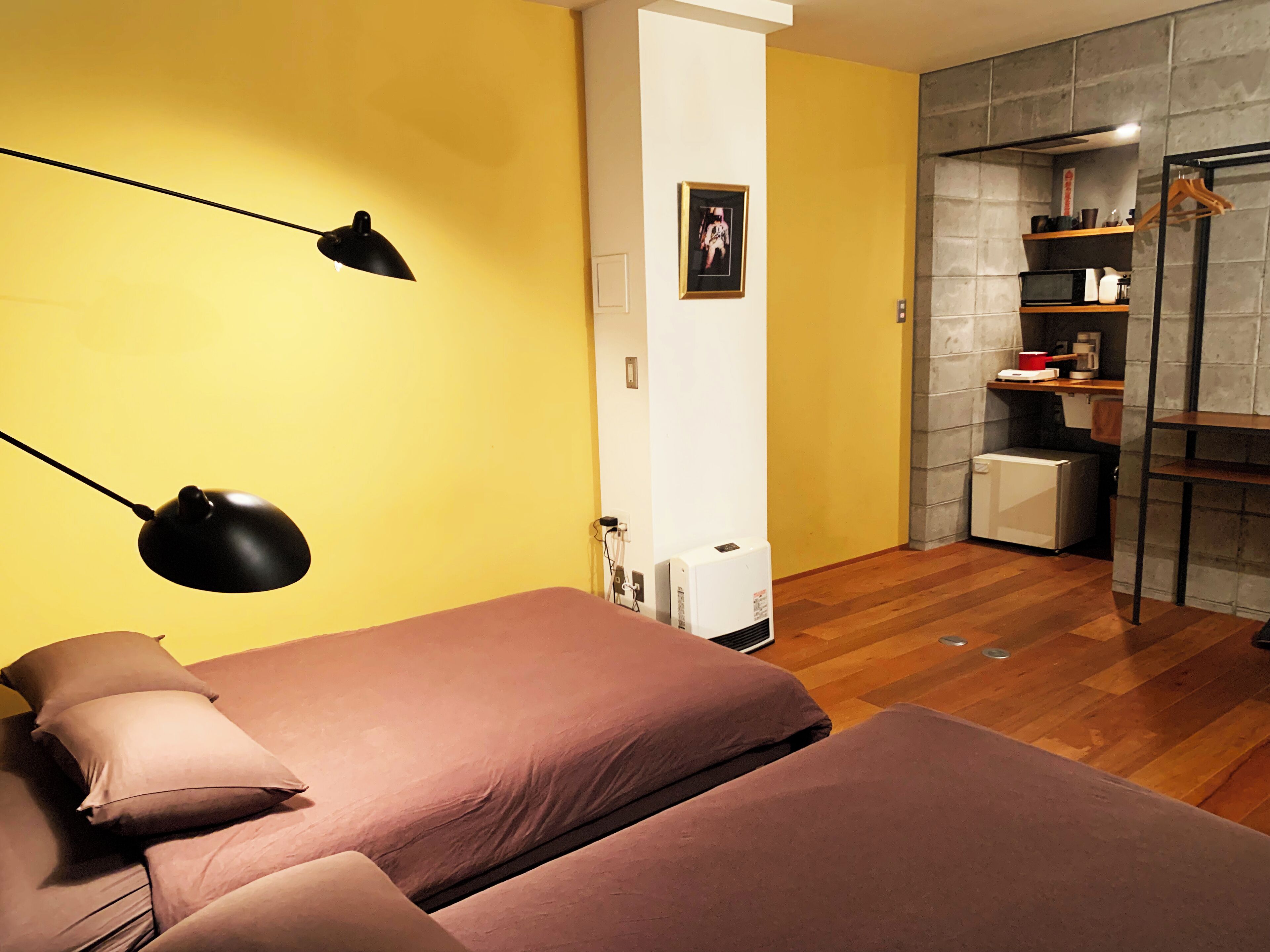 Una habitación con dos camas, un escritorio, una silla, una lámpara, un cuadro en la pared y una pequeña kitchenette.