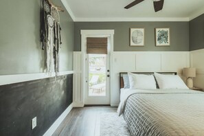 Une chambre à coucher comprenant un lit, un ventilateur de plafond, une porte donnant sur un balcon et deux œuvres d’art accrochées au mur.