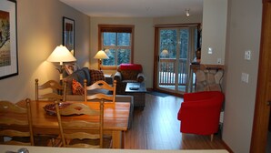A cozy living room with a dining area, wooden furniture, and a large window with a view of trees. - Convenient and located in the resort village (Sun Peaks)