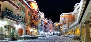 A snow-covered street lined with brightly lit buildings, suggesting a winter setting.