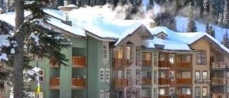 Ein mehrstöckiges Hotel mit Balkonen, umgeben von schneebedeckten Bäumen und einer Bergkulisse.