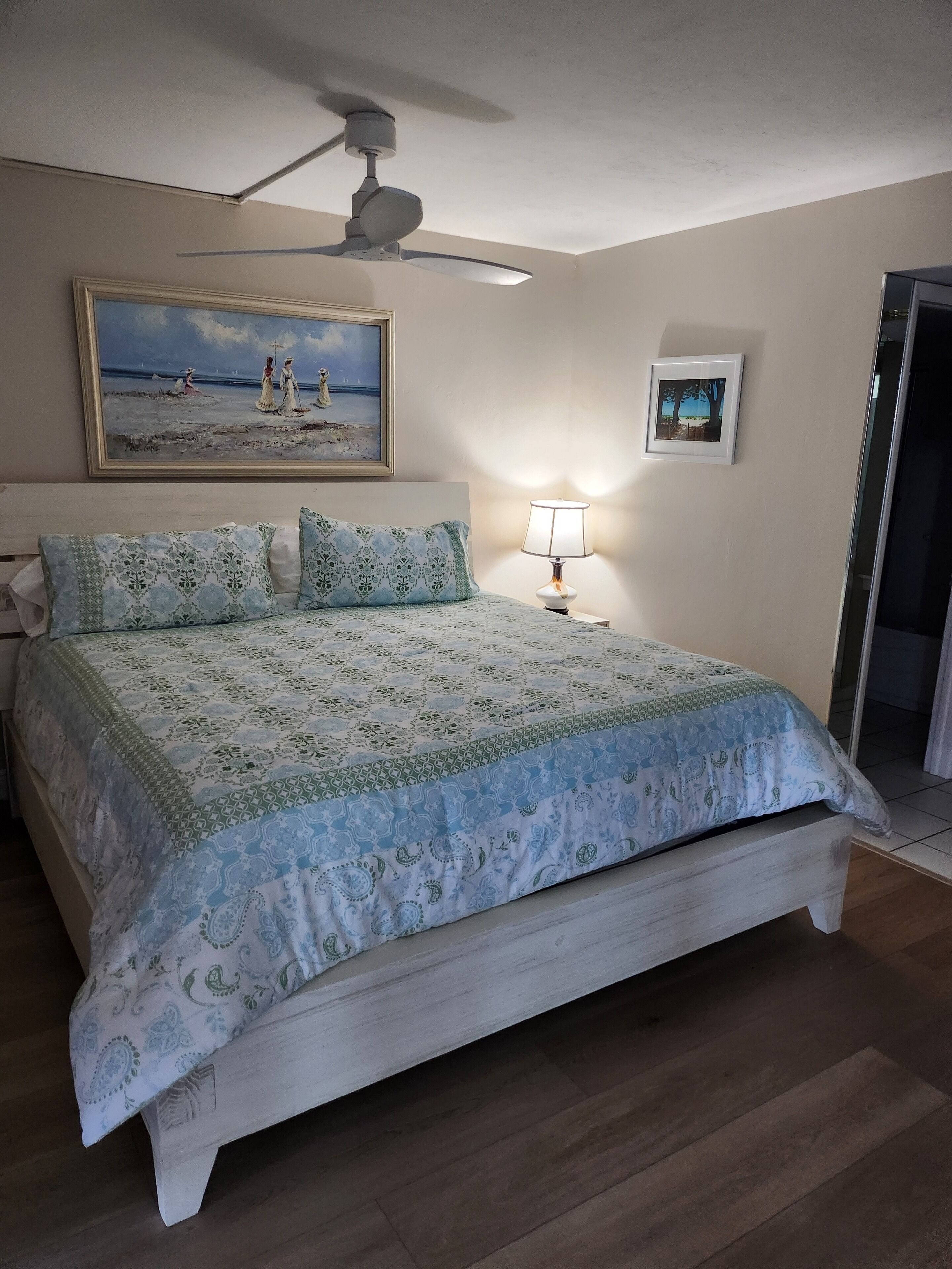 A bedroom with a bed, a ceiling fan, a painting, a nightstand with a lamp, and a framed picture on the wall.