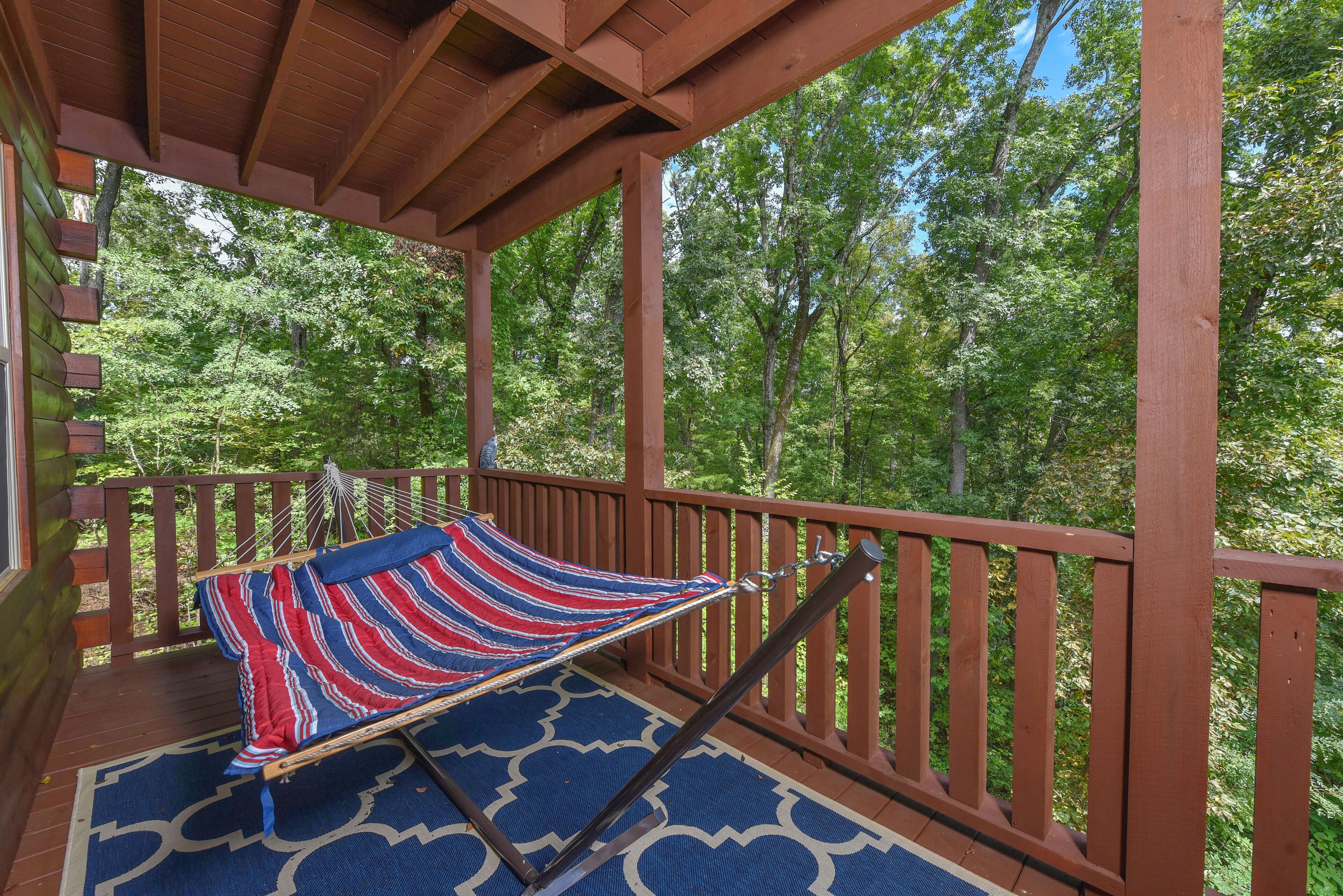 Un hamac sur une terrasse en bois, agrémenté d’un tapis à motifs, entouré d’une balustrade en bois et d’arbres.