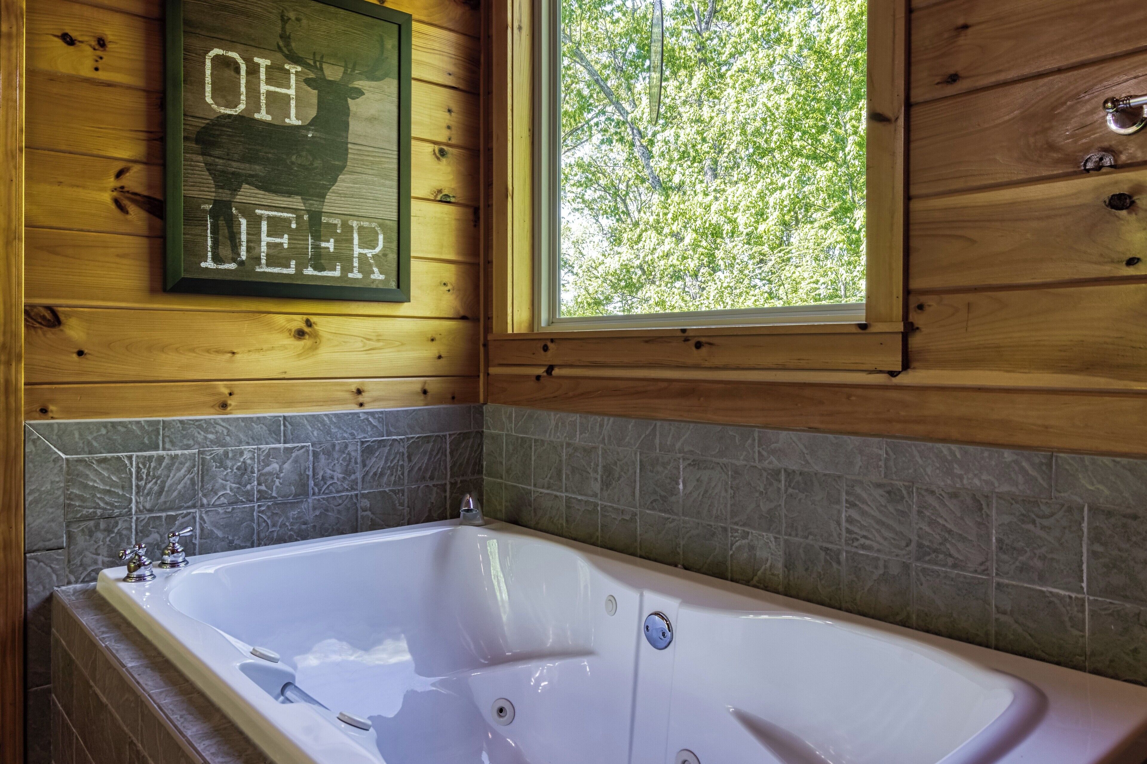 A wooden cabin bathroom with a large bathtub, a window with a view of trees, and a sign that reads "OH DEER".