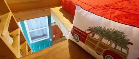 Une chambre avec un lit en mezzanine, une fenêtre et un coussin rouge et blanc orné d’un motif représentant une voiture et un sapin de Noël.