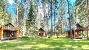 A cabin with a porch and a deck, surrounded by tall trees and a grassy area. - Swan River Retreat: 5 Cabins on River. Sleeps 30.  Family Reunions! (Bigfork)