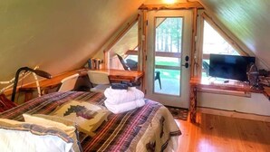 A cozy attic bedroom with a bed, a desk, and a window overlooking a green landscape. - Swan River Retreat: 5 Cabins on River. Sleeps 30.  Family Reunions! (Bigfork)