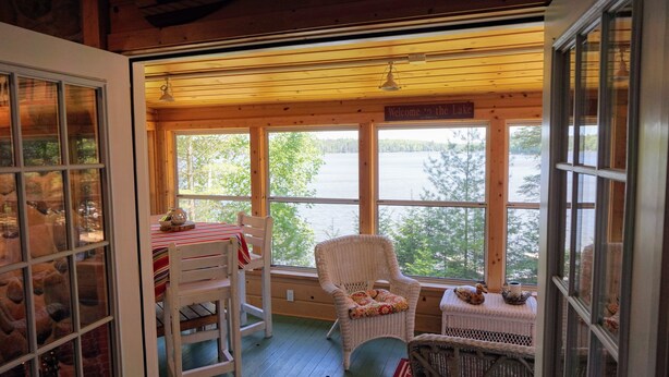 Una cabaña acogedora con un ventanal grande que da a un lago, muebles de mimbre y un letrero que dice 'Bienvenido al Lago'.