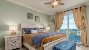 A bedroom with a bed, nightstands, a bench, a ceiling fan, and a view of the ocean through large windows.