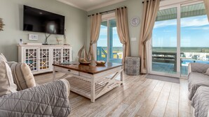 A spacious living room with a large TV, a wooden coffee table, and a view of the sea through large windows.