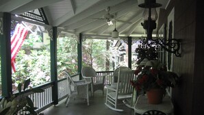 A covered porch with white wicker chairs and a rocking chair, surrounded by greenery and a view of a house. - ​Trailside Gretna Escape ▪︎ Walkable 3br Cottage  (Mt Gretna)