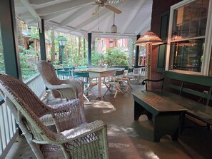A covered outdoor seating area with wicker chairs and a table, surrounded by greenery.
