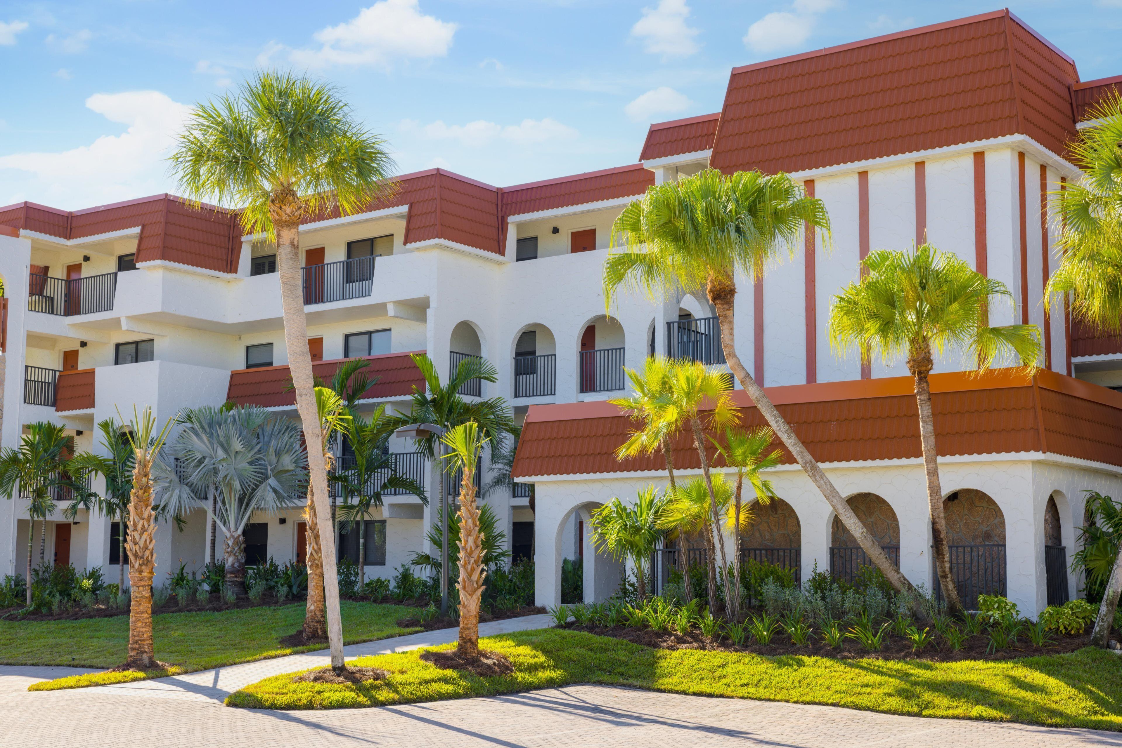 A multi-story hotel with a red roof and white walls, featuring balconies and a courtyard with palm trees.
