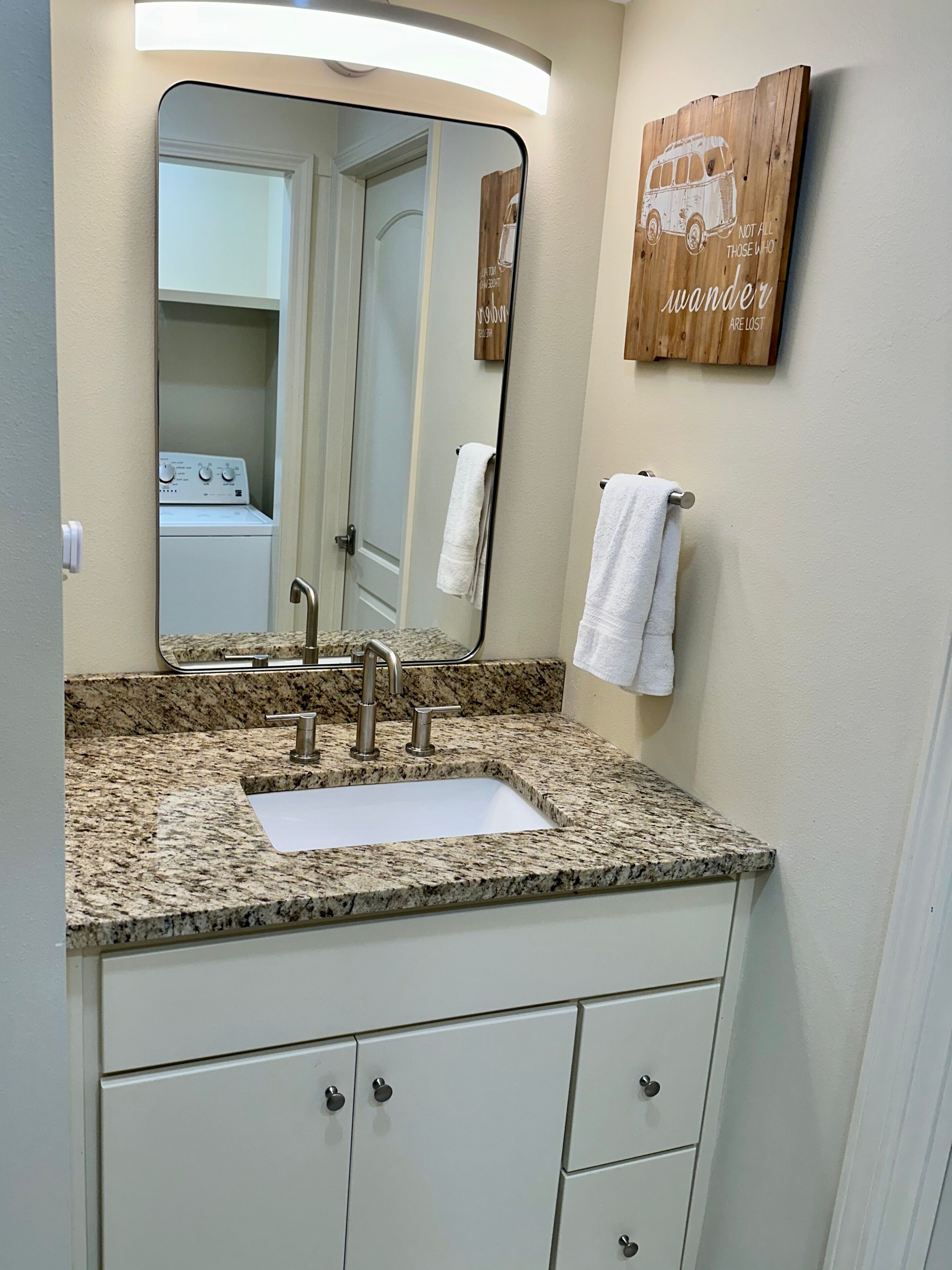 Une salle de bain avec un lavabo doté d’un plan de travail en granit, un miroir, un porte-serviettes et une plaque en bois portant l’inscription : « Tous ceux qui errent ne sont pas perdus. »