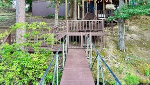 Een houten brug die naar een gebouw met een balkon leidt, omringd door bomen en een waterpartij.