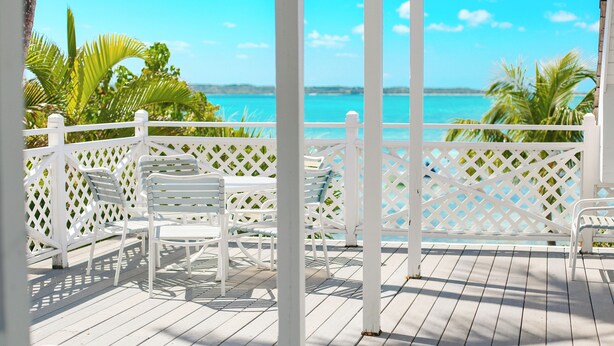 Eine Holzterrasse mit weißen Stühlen und einem weißen Zaun mit Blick auf das türkisblaue Meer.