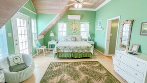 Ein Schlafzimmer mit schrägen Wänden, einem Bett mit blumigem Überwurf, einem Stuhl, einer Kommode und einem großen grünen Teppich.