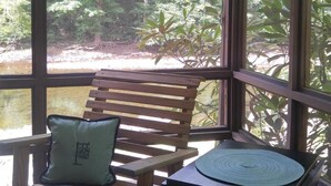 Un banco de madera con cojín y una mesa pequeña con posavasos, todo en un ambiente luminoso con grandes ventanales que dan a un bosque.