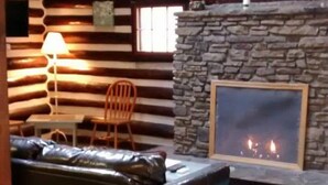 Interior de una cabaña de troncos con chimenea de piedra, un sofá de cuero y una silla de madera.