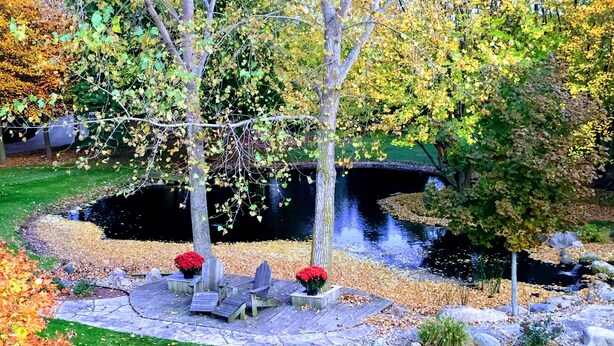 A circular stone patio with wooden chairs and red flower pots, surrounded by a grassy area and trees with autumn leaves.