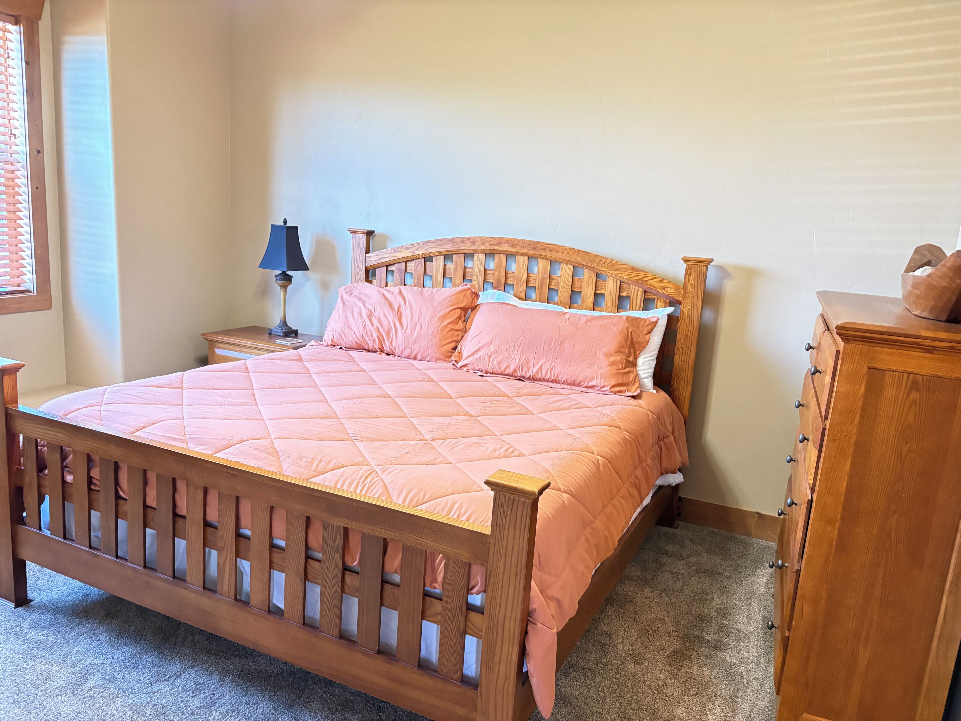 A bedroom with a wooden bed, a nightstand with a lamp, a dresser, and a window with blinds.