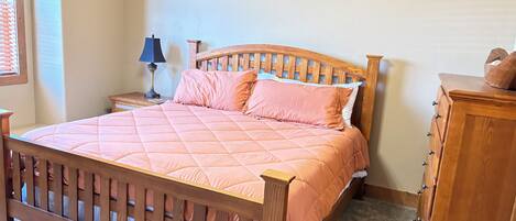 A bedroom with a wooden bed, a nightstand with a lamp, a dresser, and a window with blinds.