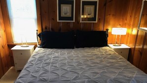 A bedroom with a large bed, two nightstands, a window with blinds, and two framed pictures on the wall. - Cabin in village of Suttons Bay! (Suttons Bay)