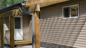 Une terrasse en bois avec une gouttière en métal, des fondations en pierre et une fenêtre sur une maison dont le revêtement extérieur est constitué de planches disposées horizontalement.